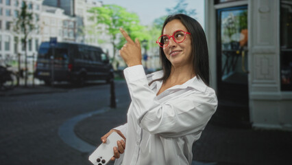 Obraz premium Young woman points finger and holds smartphone on city street wearing red glasses and white shirt while smiling broadly; playful joy.