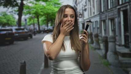 Fototapeta premium Young brunette woman holding makeup brush covers mouth with hand while smiling on a cobblestone city street lined with row houses; surprise playfulness.