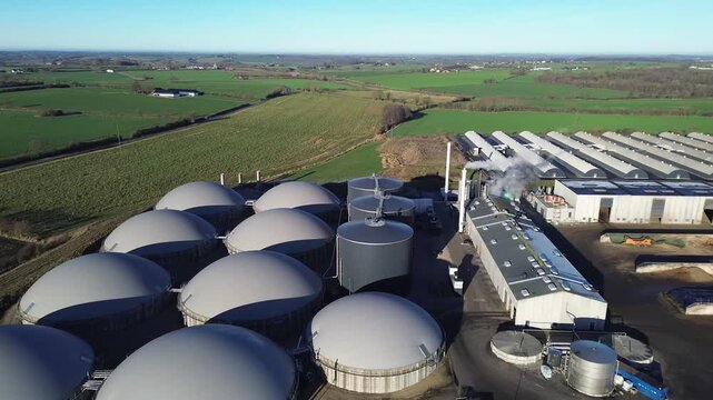 Danish biogas facility featuring large digesters, storage tanks, and processing buildings surrounded by agricultural fields. The scene highlights modern renewable energy production and sustainable was