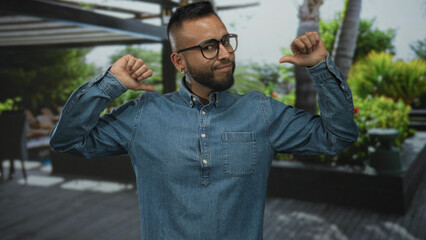 Man in denim shirt and glasses, thumbs pointing to chest by resort pool on outdoor wooden deck with palm planters and lounge chairs; confidence.