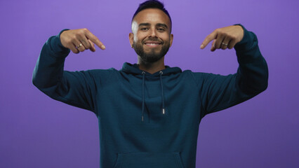Naklejka premium Young hispanic man pointing down with hands in purple studio, wearing teal hoodie and smiling; promotion confidence.