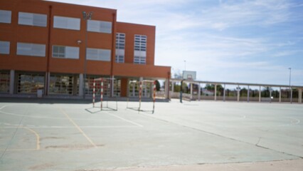 Blurred outdoor school sports court with brick building, basketball hoop and goal posts in soft defocused focus; background backplate copyspace calm.