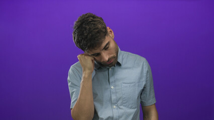 Fototapeta premium Young hispanic man in short sleeve shirt with hand on cheek supporting his head in studio setting while eyes closed; fatigue.