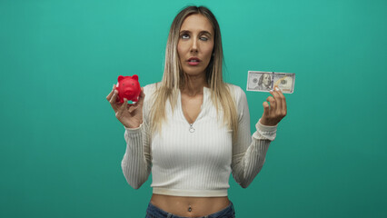 Woman holding red piggybank in left hand and holding us dollar banknote in right hand in teal studio; saving versus spending uncertainty.