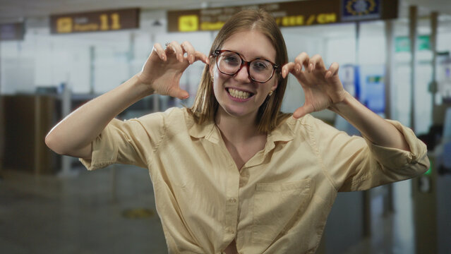 Young blonde woman with glasses raises hands in claw shape at an airport terminal checkpoint; playfulness.
