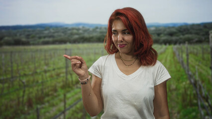 Fototapeta premium Young hispanic woman in white t shirt smiling slightly points finger to vineyard rows in forest while standing among green vines; quiet confidence.