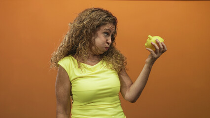 Woman inspects yellow piggy bank held in right hand, puffing cheeks and squinting face in a studio;...