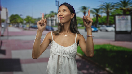 Fototapeta premium Thai woman with bare forearms points index finger upward on a sunny street with palm trees and parked cars; optimism curiosity.