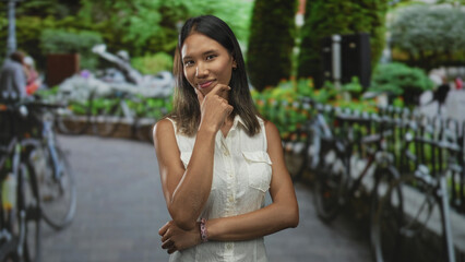 Fototapeta premium Thai woman touching chin and posing by a bicycle rack on street, wearing white sleeveless top; thoughtful contemplation.