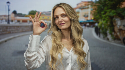 Fototapeta premium Woman smiling outdoors with blonde hair in a street setting gestures okay sign while wearing a white shirt, embodying happiness and confidence with a charming expression.
