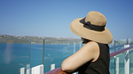 Fototapeta premium Woman smiles and leans on railing on building deck while wearing sunhat and sunglasses; serenity vacation.