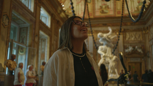 Woman looking up with visible face and glasses in ornate building gallery, wearing layered necklace and white shirt over black top; awe curiosity.