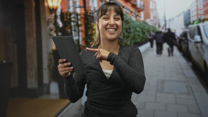 Young hispanic woman holding tablet points finger to screen on city street near storefront, smiling and engaging with device  joy engagement connection. © Krakenimages.com