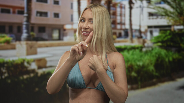 Woman in blue bikini top pointing finger to herself on sunlit street with blurred buildings and greenery in background; denial.
