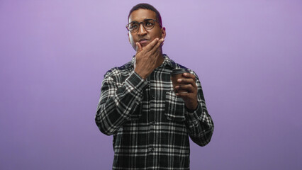Obraz premium Man in glasses and plaid shirt holding a takeaway coffee cup, hand to chin and touching cheek while smiling in studio; thoughtful calm introspection.