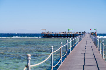 Fototapeta premium Wooden pier extends over clear turquoise water towards a seaside platform with palm trees and a blue sky in the background
