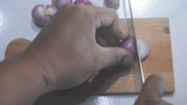 Person slicing shallots on a chopping board with a knife. Simple food prep scene showing fresh vegetables, spice ingredients, and traditional cooking process.