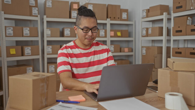 Man puts on glasses and types on laptop at a packing desk surrounded by stacked parcels and shelving in building; focused productivity.