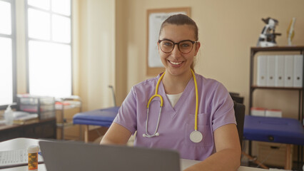 Woman doctor in clinic wearing purple scrubs and yellow stethoscope, typing on laptop while reviewing a prescription bottle on desk; focused caring.
