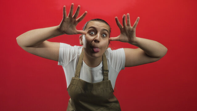Woman in apron sticks tongue out and frames eyes with hands while posing against a red studio backdrop; playful mischief.