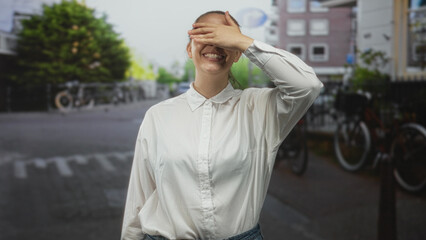 Fototapeta premium Woman covering eyes with her hand while smiling in a white shirt on a city street with parked bicycles; playful surprise.