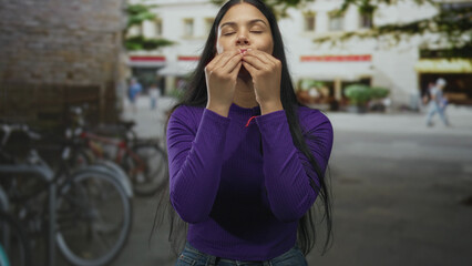 Fototapeta premium Woman in purple sweater pinches fingertips to lips in sunlit city street by parked bicycles; delight taste flavor satisfaction.