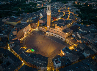 Obraz premium Aerial night view of Torre del Mangia rising above Piazza del Campo in Siena, Tuscany. Illuminated medieval tower and historic city center seen from above during evening hours