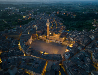 Obraz premium Aerial night view of Torre del Mangia rising above Piazza del Campo in Siena, Tuscany. Illuminated medieval tower and historic city center seen from above during evening hours