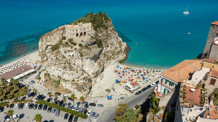 Aerial view of Sanctuary of Santa Maria dell'Isola in Tropea, Calabria, Italy. The ancient church stands on a rocky promontory, overlooking a crowded sandy beach filled with colorful sun umbrellas. © Stefano Tammaro