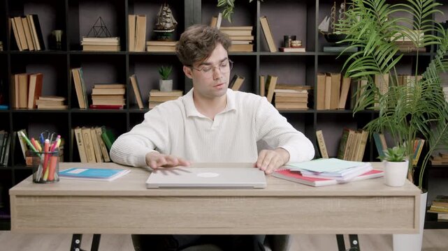 Young male student in white sweater organizes study materials on a desk, adjusting papers and stationery in a well-lit home office with bookshelves in the background