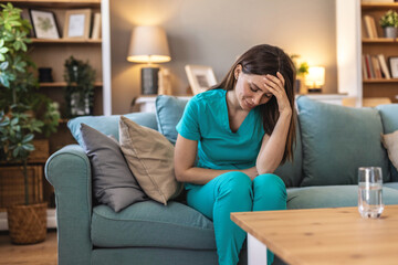 Obraz premium A nurse wearing scrubs is sitting in an armchair, appearing exhausted after a long shift. The setting is a peaceful home environment with natural light filtering through the window. 