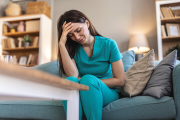 Obraz premium A healthcare professional in scrubs sits on a couch, appearing stressed and thoughtful. The image captures a moment of reflection in a relaxed home setting. Ideal for concepts of burnout and self-care