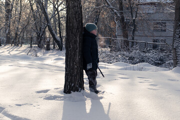 A little boy stands next to a tree on a cold winter day in a snowy park