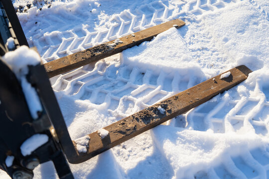 Forklift style pallet fork attachment in snow with two steel tines and worn rust patches, bright sunlight highlighting tire track pattern and tool wear