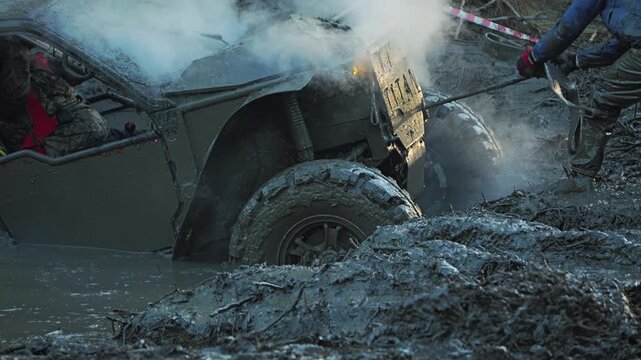 Extreme off-road vehicle stuck deep in a mud bog during a competition. Steam rises from the hot engine as a person pulls a tow rope to assist. Gritty, action-packed scene of motorsport. Slow motion