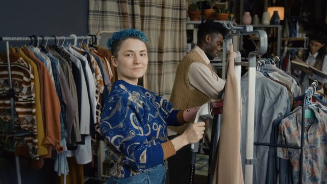 Slow motion portrait shot of young woman with short curly blue hair and prosthetic arm smiling for camera while steaming jacket in thrift store