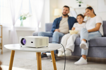 Obraz premium Home projector ready for happy family watching movie. Digital device on table in living room, parents and child relax on sofa with popcorn and remote, people enjoying cozy TV together at weekend