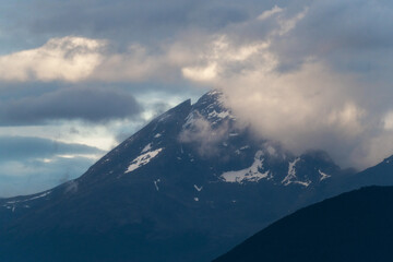 Dramatic view of a rugged, snow-dusted mountain peak in Tierra del Fuego along the Beagle Channel, shrouded in moody, heavy clouds. The atmosphere is cold and remote Patagonian wilderness.