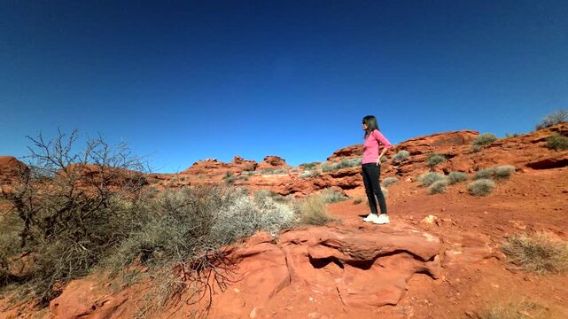 Looking up at woman standing on rugged terrain near vibrant red boulders of  sandstone formation deep blue sky background 60FPS 4k