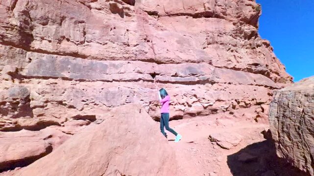 Woman hiking along base of unique boulder formation of red sandstone and rocky cliff landscape with deep blue sky