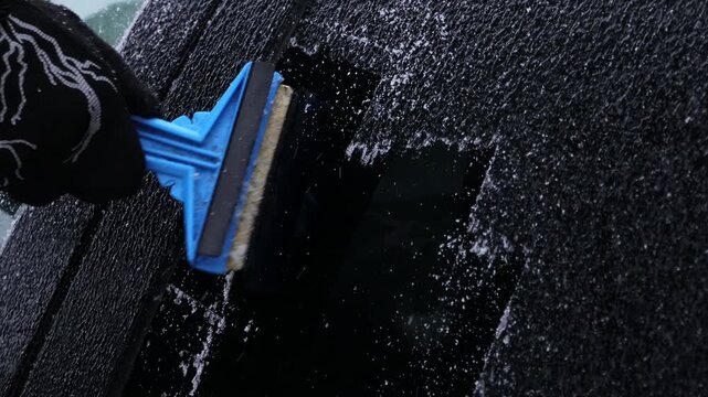 Hand using blue ice scraper to remove ice from car windshield, revealing clear glass underneath, with ice shards scattered on the surface and snow in the background