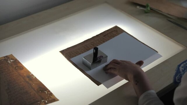 A professional conservator begins the first steps of the restoration process, carefully aligning and stabilizing a historical manuscript page using weights on a light table.