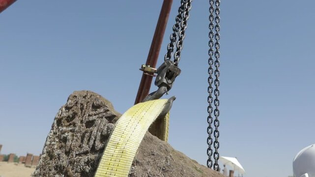 During a restoration project, a large carved stone block is carefully lifted using a yellow lifting sling and a manual chain hoist system.