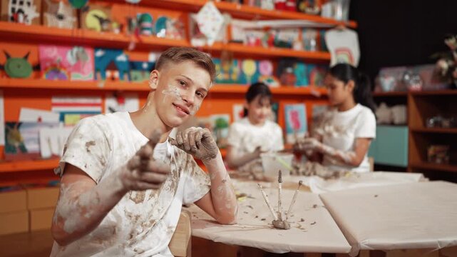 high school boy looking at camera while multicultural children modeling clay. Diverse student having pottery class together. Child smile while wearing white shirt with mud stained cloth. Edification