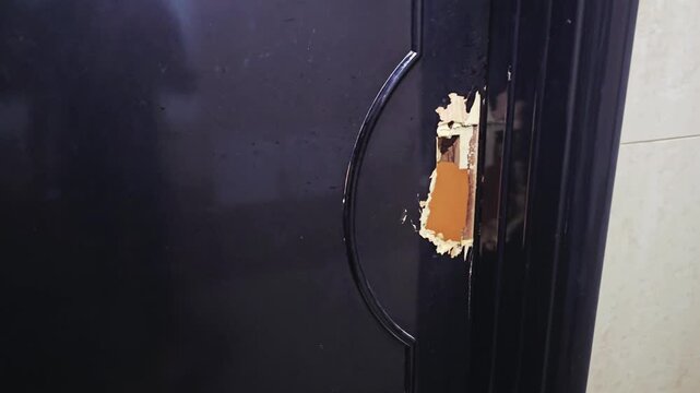 Damaged black main entrance door with a broken lock mechanism and curved handle, showing signs of forced entry or wear.