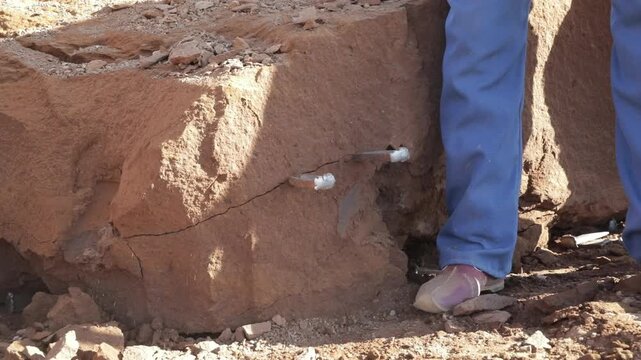 A stonemason using a sledgehammer to strike steel wedges to create a crack and split a massive rock block.