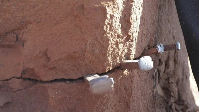 A worker using a sledgehammer and metal chisels to pry and extract large rock pieces from the ground using traditional hand tools.