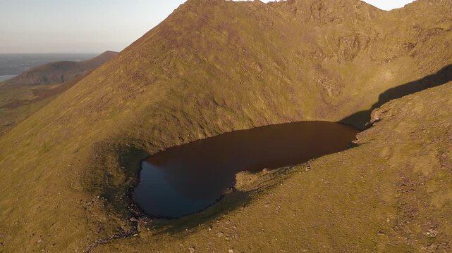 Aerial view shows a tranquil corrie lake high on Carrauntoohil mountain in County Kerry Ireland. The sun casts long shadows and warm light across the rugged terrain during late afternoon.