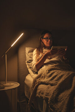 Woman reading on a digital tablet in bed at night with a modern lamp