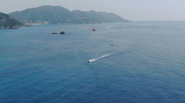 Aerial wide shot of vast blue sea with boat towing parasailer and misty mountains on horizon during sunny afternoon